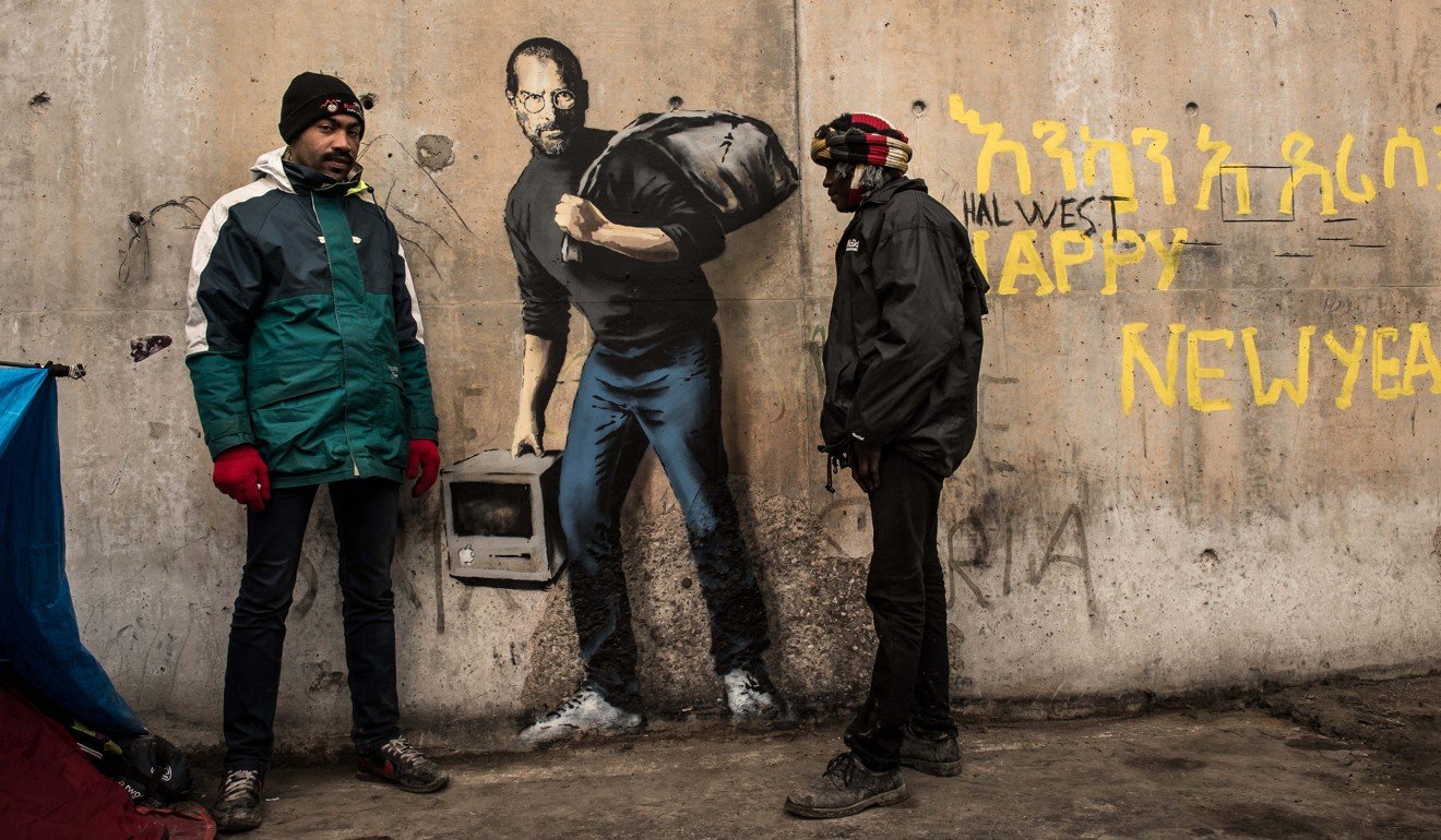 Two men stand next to a street art graffiti by elusive British artist Banksy representing Steve Jobs, founder and late CEO of Apple, at the migrant camp known as the “Jungle” in Calais. Photo: AFP/ Banksy